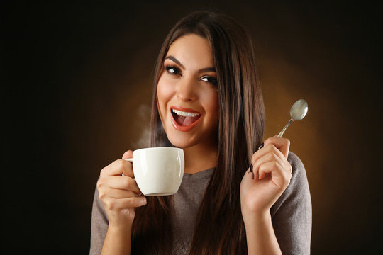 Happy beautiful woman with cup of coffee and spoon on dark background