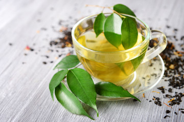Glass cup of tea with green leaves and scattered tea around on grey wooden background
