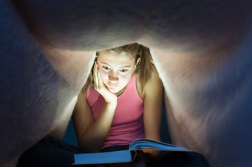 Young girl hiding under blanket and enrapt reading interesting b