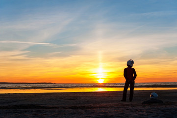 Female biker silhouette standing on multicolored sunset. Blue sk
