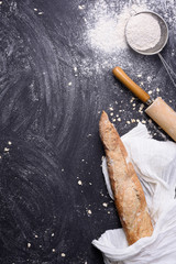 French baguette or rustic bread wrapped in white towel with rolling pin and flour over black background. Top view, copy space.