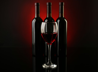 Wine glass against bottles in a row on dark red background, close up