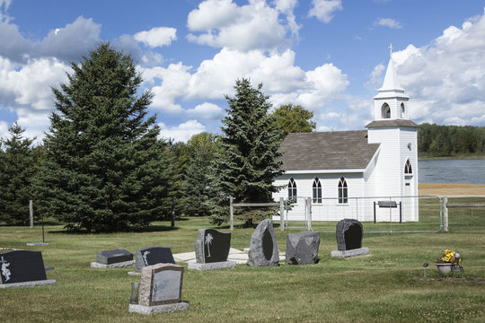 horizontal image of a little white country church sitting around  a grave yard surrounded by green trees under a beautiful blue sky with white puffy clouds.