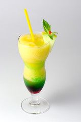 descending close-up of a hurricane glass filled with a layered fruit cocktail, with purpple and green syrup on the bottom, decorated with a melon wedge and a yellow straw, on a gray background