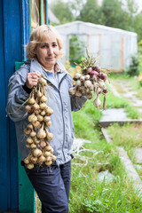A middle-aged woman stands near the house and holds a bunch of onions and garlic in her hands.
