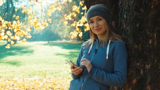 Young adult woman using smartphone smiling at camera standing near tree in the park in the autumn