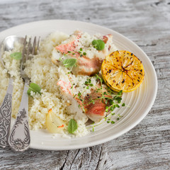 Baked fish with lemon and herbs and cous cous on a white plate, on bright wooden surface. Healthy food