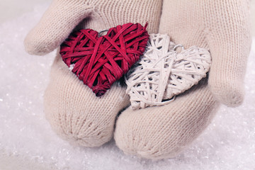 Female hands in white knitted cozy mittens holding two hearts   on a white snow background. Christmas tree decoration. Love concept. Winter and Christmas time
