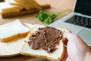 A man holding a bread with chocolate spread while working with laptop in the morning. Focused on a sliced of bread with chocolate spread.