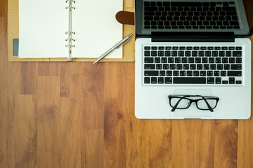 Wooden table with laptop, notebook, pen and eye glasses on top. Top view with copy space.