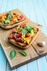 Fresh homemade rustic bruschetta with chopped red, yellow and green tomatoes, basil, olive oil and toasted garlic italian bread ciabatta on wooden cutting board over turquoise background close-up