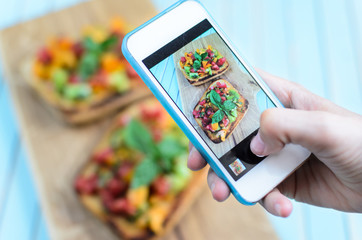 Taking photo on smartphone of fresh homemade rustic bruschetta with chopped tomatoes, basil and olive oil on toasted garlic bread on wooden board over turquoise background