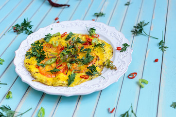 Delicious breakfast omelette or scrambled eggs with red and yellow tomatoes, red hot chili peppers and parsley on patterned plate over stylized old aged turquoise table background close-up, copy space
