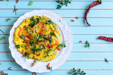 Delicious breakfast omelette or scrambled eggs with red and yellow tomatoes, parsley and dried red hot chili peppers on patterned plate on old aged turquoise table background, top view with copy space