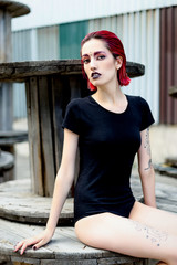 Attractive young woman with colorful makeup and red hairstyle in simple black bodywear sitting on big wooden reels