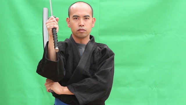 An Asian man in a Japanese Gi draws a Samurai Sword (Katana), and slashes through the air with sword and scabbard (siniwali style), then resheaths his blade in front of a green screen.
