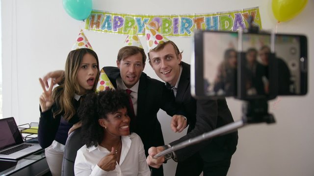 Business woman celebrating her birthday and doing a party with colleagues in her office. A friend holds his phone with a selfie stick and takes pictures of his fellow coworkers smiling. Medium shot