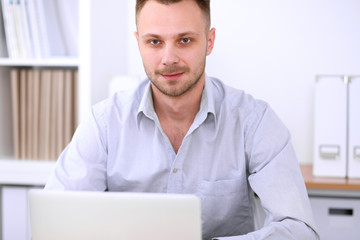 Portrait of young business man sitting at the desk on office background.