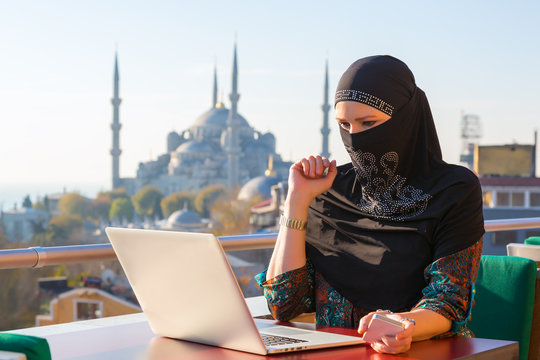 Traditionally dressed Muslim Woman working on computer at outdoor balcony with oriental urban landscape on background
