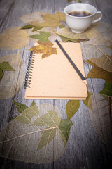 coffee and notebook on wooden table surrounded by dry leaves