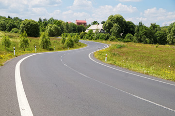 Empty asphalt road with a double bend between flower meadows in the countryside. Village among the green leafy trees background. Clear sunny day with blue skies and white clouds.