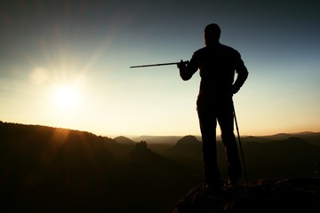 Tourist guide show the right way with pole in hand. Hiker with sporty backpack stand on rocky view point above misty valley. Sunny spring daybreak in rocky mountains.