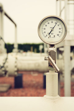 Closeup of a high pressure manometer, measuring natural gas pressure. Pipes and valves in the background. Selective focus.