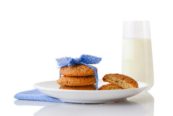 Stack of three homemade oatmeal cookies tied with blue ribbon in small white polka dots and halves of cookies on white ceramic plate on blue napkin and glass of milk, isolated on white background