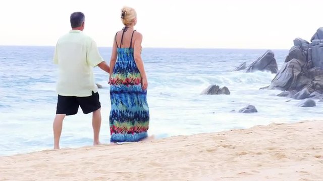 An older couple holding hands and walking down the beach in front of large rocks