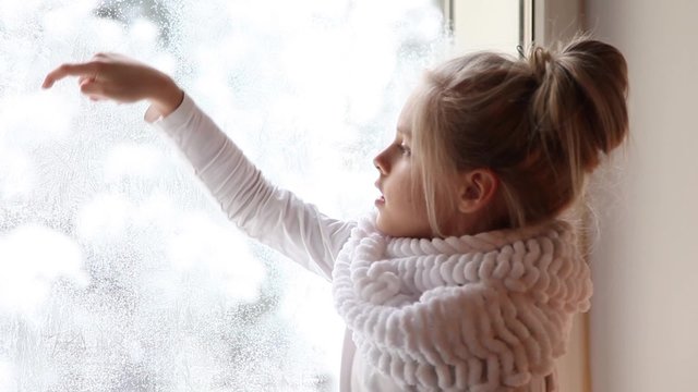 Красивая, милая девочка рисует рисунок на заснеженном окне.