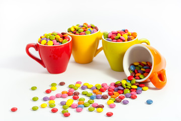 Plenty assorted color dragees in four small bright colored cups, the orange cup is felt over the sweets are spilled over the white table