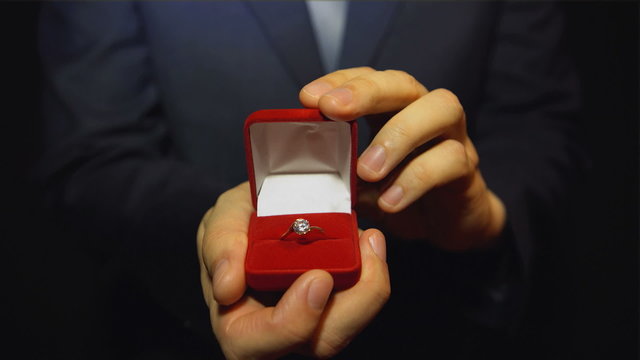 A young man in the blue suit giving a ring with a diamond in the red box
