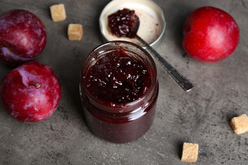 A jar of tasty jam, a spoon, plums and crackers on grey background