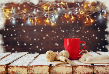 Obraz na płótnie Canvas Cup of hot coffee and cozy knitted scarf on wooden table in front of garland lights background 