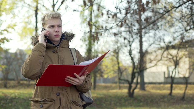 Boy talking on cellphone and holding red binder in the autumnal park
