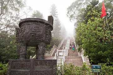 Handful of people climbing long flight of stairs to the Tian Tan Buddha or Big Buddha statue in Hong Kong, China, at a foggy morning.