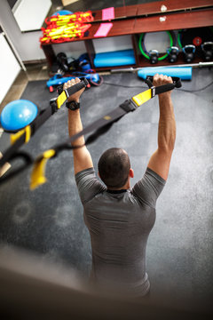 A determined male gym member focuses on his fitness goals as he works out with intensity on a TRX suspension trainer, targeting various muscle groups to stay strong and fit.