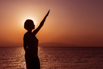 Fototapeta premium Silhouette of young girl standing on the beach with hand up, at the sunset against the sun