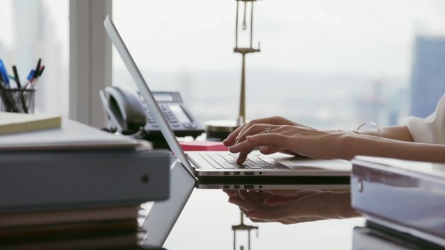 Adult businesswoman sitting in modern office with beautiful sight of the city. The secretary writes fast on laptop pc. Closeup shot of hands and keyboard