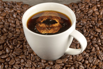 Weiße Tasse mit Kaffee gefüllt, mit Kussmund verziert steht in einem Meer von Kaffeebohnen