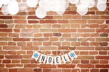 Black and white NOEL and blue snowflake paper card homemade flag garland with top blurred lights border decoration on rustic brick wall background. Horizontal postcard with space for text or lettering