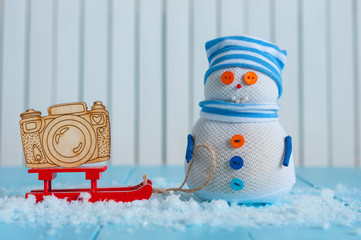 Handmade snowman in striped cap with red sled and toy camera on snow In winter. Christmas postcard...