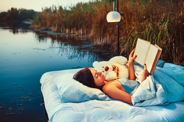 Pretty woman is laying on the floating bed with a book and toy.
