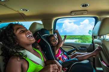 Brazilian girls singing and laughing sitting on backseats in car. Travelling female children in child safety seat with seatbelt on a summer day in tropical area - 97593500