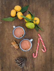 Christmas or New Year attributes. Fresh mandarins with leaves, cinnamon sticks, pine cone, hot chocolate in mugs and candy canes over rustic wooden background, top view