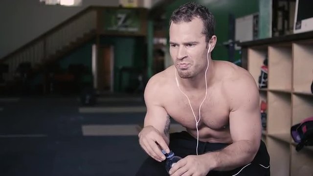 A fit young man sitting in a gym takes a drink out of a water bottle and then looks at the camera
