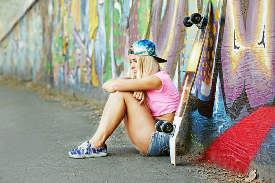Young woman with skating board sitting on asphalt on painted wall background