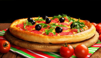 Tasty pizza with salami and olives decorated with tomatoes on wooden table against black background