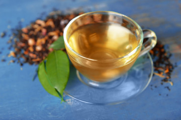 Glass cup of tea with green leaves and scattered tea around on blue wooden background