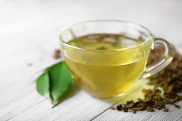 Glass cup of tea with green leaves and scattered tea around on grey wooden background, close up